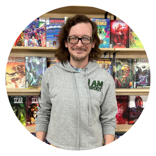 Staff member Blake, a petite man with long wavy brown hair with well-groomed facial hair, a grey sweatshirt with text that reads "I am groot", standing in front of the comic book wall